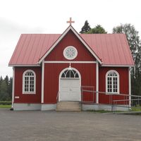 Litet kapell, rött hus med vita knutar, dörr i mitten med inva-ramp till höger, parkeringsplats framför byggnaden