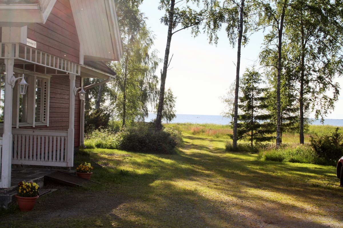 sommarbild med blick en ut mot havet. Huvudbyggnadens ingång till vänster och på mitten går en väg mot havet, på båda sidor om vägen står några björkar.